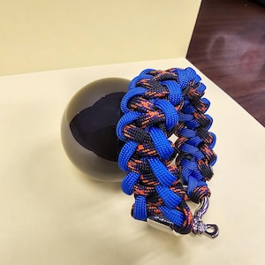 Può includere: Un braccialetto in paracord blu e nero con accenti arancioni e una chiusura argentata. Il braccialetto è realizzato con un motivo intrecciato ed è uno stile popolare per gli appassionati di attività all'aperto.