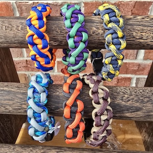 May include: Six colorful paracord bracelets with different patterns and colors, including orange, blue, green, purple, gray, black, and brown. The bracelets are displayed on a wooden surface.