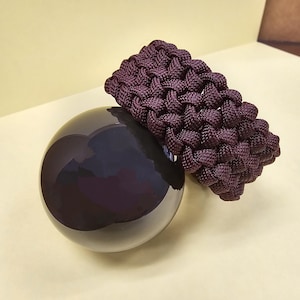 May include: A dark purple, round, glass orb with a dark purple paracord bracelet wrapped around it.