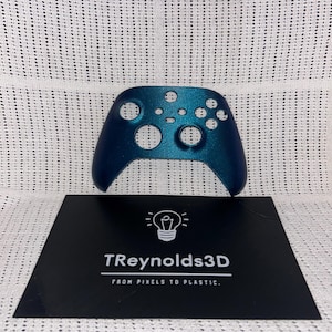 May include: A teal-coloured video game controller is positioned above a black business card. The controller features several holes and buttons. The business card displays the text "T Reynolds3D FROM PIXELS TO PLASTIC" and a lightbulb graphic.