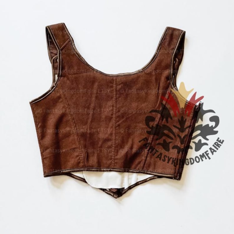 May include: A sleeveless brown bodice with a scoop neckline and structured seams. The garment features a pointed hemline, a side zipper closure, and a white interior lining. A dark watermark logo is visible on the right side of the brown fabric.
