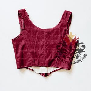 May include: A burgundy velvet corset top with a scoop neckline and shoulder straps. The bodice features vertical seams and a decorative panel on the side. The lining is white. The corset is designed for a Renaissance or fantasy aesthetic.