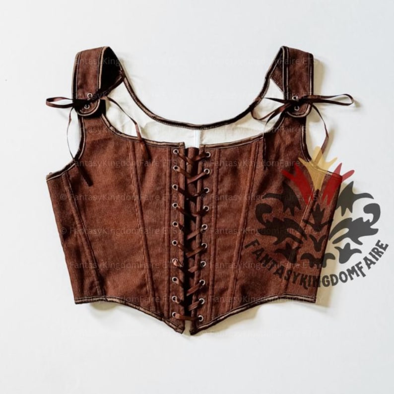 May include: A brown sleeveless bodice featuring a front lace-up closure with dark brown ribbons and metal eyelets. The garment has wide shoulder straps with ribbon ties and a structured, fitted silhouette with vertical seams throughout the piece.
