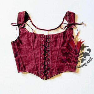 May include: A burgundy corset with a lace-up front and shoulder straps. The corset has a square neckline and is made of a textured fabric. The straps are tied with ribbons. The corset is a piece of historical or fantasy clothing.