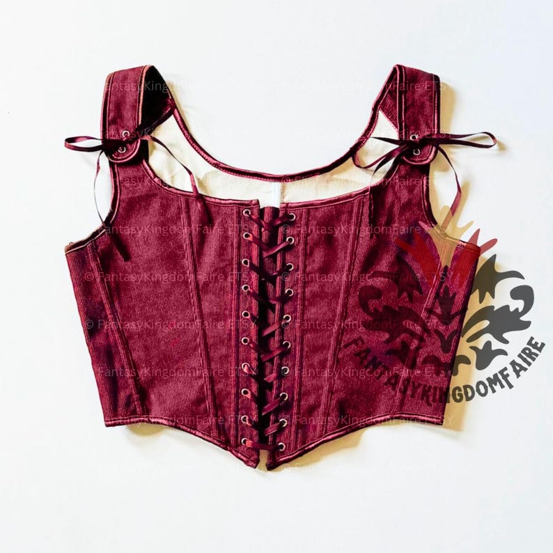 May include: A burgundy corset with a lace-up front and shoulder straps. The corset has a square neckline and is made of a textured fabric. The straps are tied with ribbons. The corset is a piece of historical or fantasy clothing.
