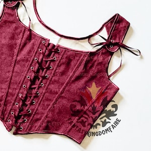May include: Burgundy corset with a lace-up front and shoulder straps. The bodice features a decorative design and is made of a textured fabric. The corset has a scoop neckline and is tied with burgundy ribbons.