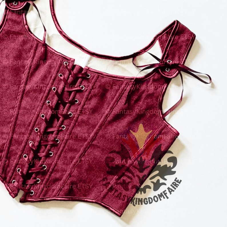 May include: Burgundy corset with a lace-up front and shoulder straps. The bodice features a decorative design and is made of a textured fabric. The corset has a scoop neckline and is tied with burgundy ribbons.