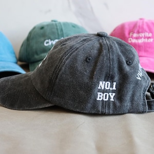 May include: A collection of baseball caps in various colors, including blue, green, pink, and gray. The gray cap in the foreground has "NO. 1 BOY" embroidered on the back. Other caps feature text and a dinosaur.