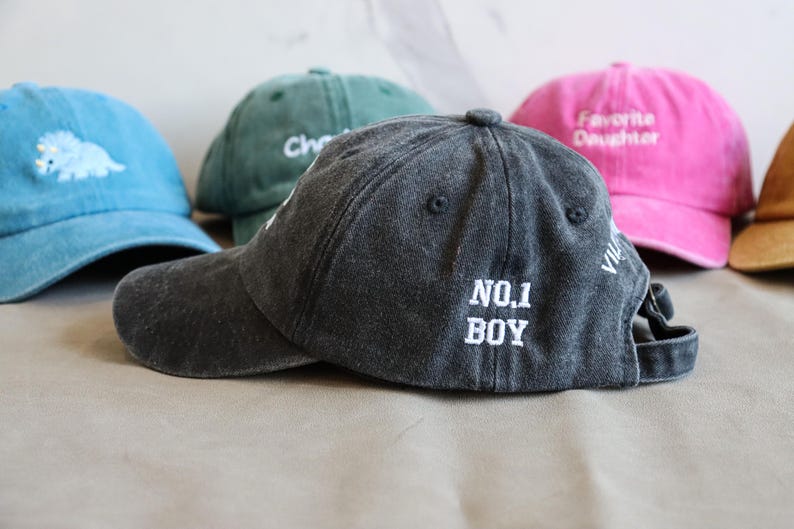 May include: A collection of baseball caps in various colors, including blue, green, pink, and gray. The gray cap in the foreground has "NO. 1 BOY" embroidered on the back. Other caps feature text and a dinosaur.