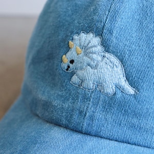 May include: Light blue denim baseball cap featuring an embroidered light blue triceratops dinosaur with yellow accents. The cap has a curved brim and a classic six-panel design, suitable for casual wear.