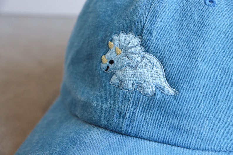 May include: Light blue denim baseball cap featuring an embroidered light blue triceratops dinosaur with yellow accents. The cap has a curved brim and a classic six-panel design, suitable for casual wear.