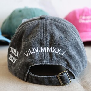 May include: A dark gray baseball cap with white embroidered text reading "NO. 1 BOY" and "VII.IV.MMXXV". Other caps in blue, green, and pink are visible in the background. The cap has a metal buckle closure.