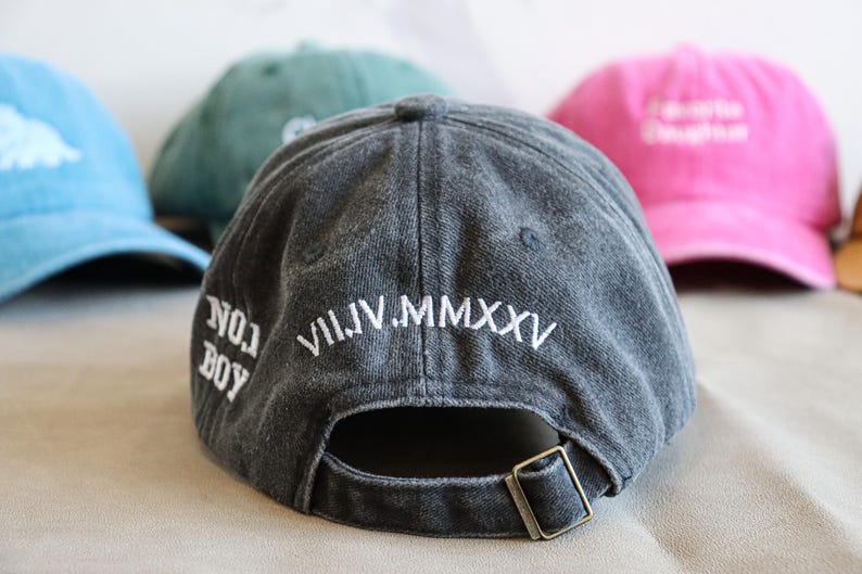 May include: A dark gray baseball cap with white embroidered text reading "NO. 1 BOY" and "VII.IV.MMXXV". Other caps in blue, green, and pink are visible in the background. The cap has a metal buckle closure.