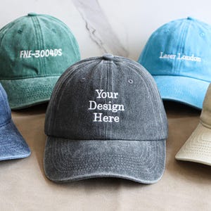 Puede incluir: Una colección de cinco gorras de béisbol en varios colores, incluyendo azul vaquero, verde azulado, azul claro, gris y blanco roto. Cada gorra presenta texto bordado, como "BEST. DAD. EVER.", "FNE-3004DS", "Your Design Here", "Laser London" y "COOL AUNT ENERGY".