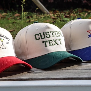 Könnte beinhalten: Drei Baseballkappen mit verschiedenen gestickten Designs. Die erste Kappe ist weiß mit rotem Besatz und dem Text "Espresso Martini Est 1995 good time" mit zwei Martini-Gläsern. Die zweite Kappe ist weiß mit grünem Besatz und dem Text "CUSTOM TEXT". Die dritte Kappe ist weiß mit blauem Besatz und einem Fußball mit einem Stern und dem Text "Team Name 2023".
