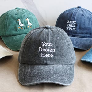 Könnte beinhalten: Fünf Baseballkappen in verschiedenen Farben, jede mit einzigartigen gestickten Designs. Die Kappen umfassen eine beige Kappe mit einem Bierkrug, eine grüne Kappe mit zwei Enten, eine graue Kappe mit "Your Design Here", eine blaue Kappe mit "BEST DAD EVER" und eine hellblaue Kappe mit einem Bären.