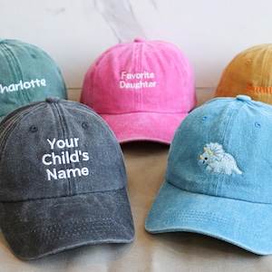 Könnte beinhalten: Fünf Baseballkappen in verschiedenen Farben: Türkis, Pink, Senfgelb, Jeansblau und Anthrazitgrau. Jede Kappe hat einen gestickten Text oder ein Design, wie einen Dinosaurier oder einen Namen.