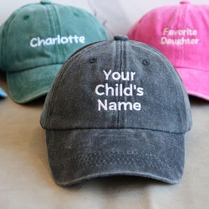 May include: A collection of personalized baseball caps in various colors, including blue, green, pink, and gray. The caps feature embroidered text such as "Charlotte", "Favorite Daughter", and "Your Child's Name". The hats are made of a soft, worn fabric.