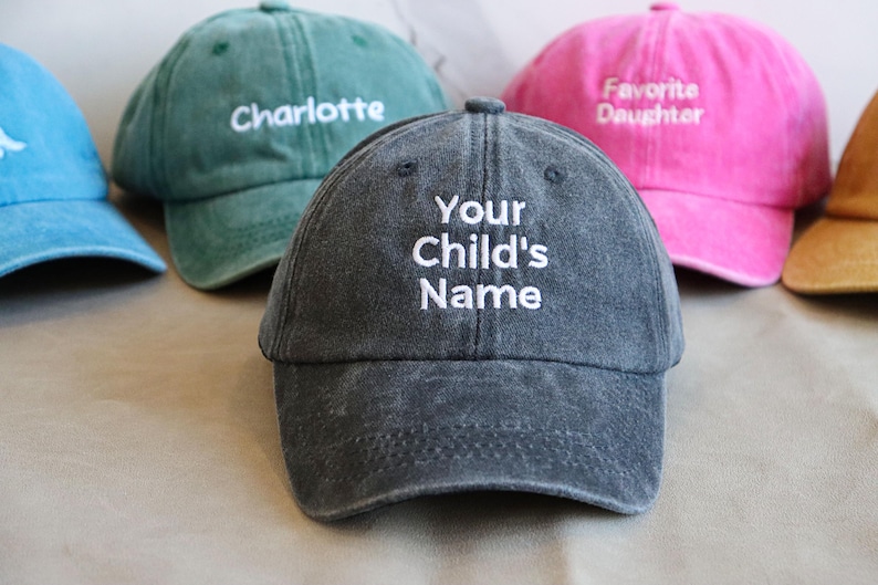 May include: A collection of personalized baseball caps in various colors, including blue, green, pink, and gray. The caps feature embroidered text such as "Charlotte", "Favorite Daughter", and "Your Child's Name". The hats are made of a soft, worn fabric.