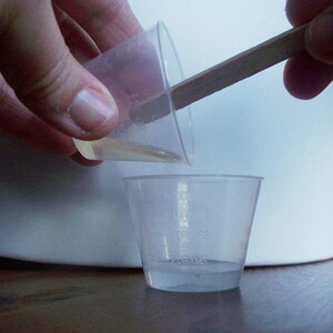 Puede incluir: Una persona vierte un líquido transparente de un vaso de plástico pequeño a un vaso de plástico más grande usando un palo de madera.