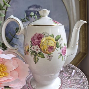 Puede incluir: Una tetera de porcelana blanca con un estampado floral de rosas amarillas y rosas con hojas verdes. La tetera tiene una tapa a juego con un pequeño pomo, un asa curva y un pico, todo ello decorado con un fino borde de color dorado.