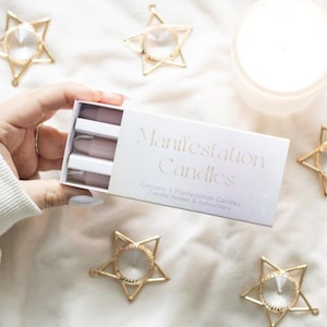 Puede incluir: Una caja blanca de "Manifestation Candles" se sostiene abierta, revelando tres velas lavanda. La caja está rodeada de adornos de estrellas doradas con centros de cristal transparente y una vela encendida. El texto de la caja dice "Contiene 3 Velas de Manifestación, Portavelas e Instrucciones."