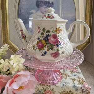 May include: A white ceramic teapot with a hexagonal shape, featuring a raised lattice texture and a colourful floral pattern of pink roses and blue flowers. The teapot has a matching lid and is displayed on a pink glass pedestal cake stand.