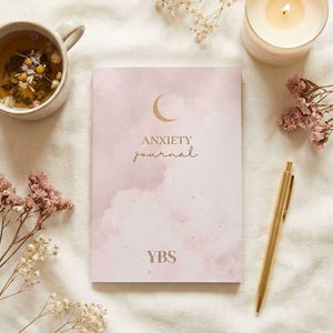 Puede incluir: Un diario rosa con las palabras "ANXIETY JOURNAL" y un diseño de media luna. Un bolígrafo dorado, una taza de té, una vela encendida y flores secas están dispuestas alrededor del diario sobre una superficie blanca.