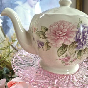 Può includere: Una teiera in ceramica color crema con un motivo floreale di fiori rosa e viola e foglie verdi. La teiera presenta un beccuccio curvo, un coperchio con pomello e un bordo smerlato, ed è posizionata su un piedistallo in vetro pressato rosa.