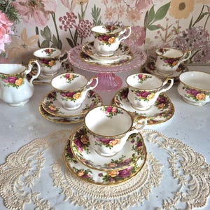 Op de afbeelding: Een set witte porseleinen theekopjes en schoteltjes met een bloemmotief van rode en gele rozen, groene bladeren en een gouden rand. De set bevat een melkkannetje, een suikerpot en een taartplateau van roze glas.