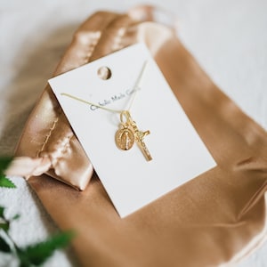 May include: Gold-colored necklace with a pendant featuring a cross and a medallion with a religious figure. The necklace is displayed on a white card with the text "Catholic Made Goods" and rests on a gold satin pouch.