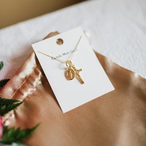 May include: A gold-colored necklace with a pendant featuring a cross and a religious medallion. The necklace is displayed on a white card with the text "Catholic Made Goods". The card rests on a gold-colored satin surface.