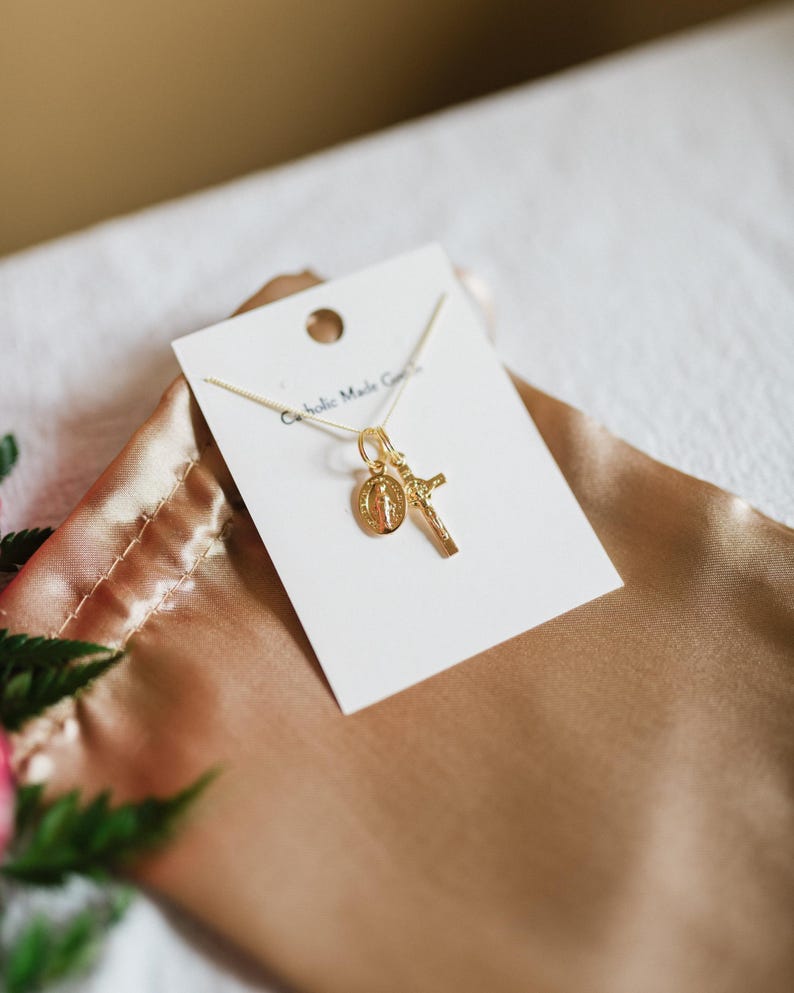 May include: A gold-colored necklace with a pendant featuring a cross and a religious medallion. The necklace is displayed on a white card with the text "Catholic Made Goods". The card rests on a gold-colored satin surface.