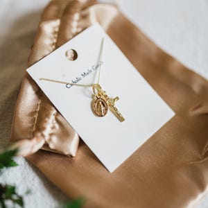 May include: A gold-colored necklace with two pendants: a cross and a medallion featuring a religious figure. The necklace is displayed on a white card inside a gold satin pouch. The card reads "Catholic Made Goods".