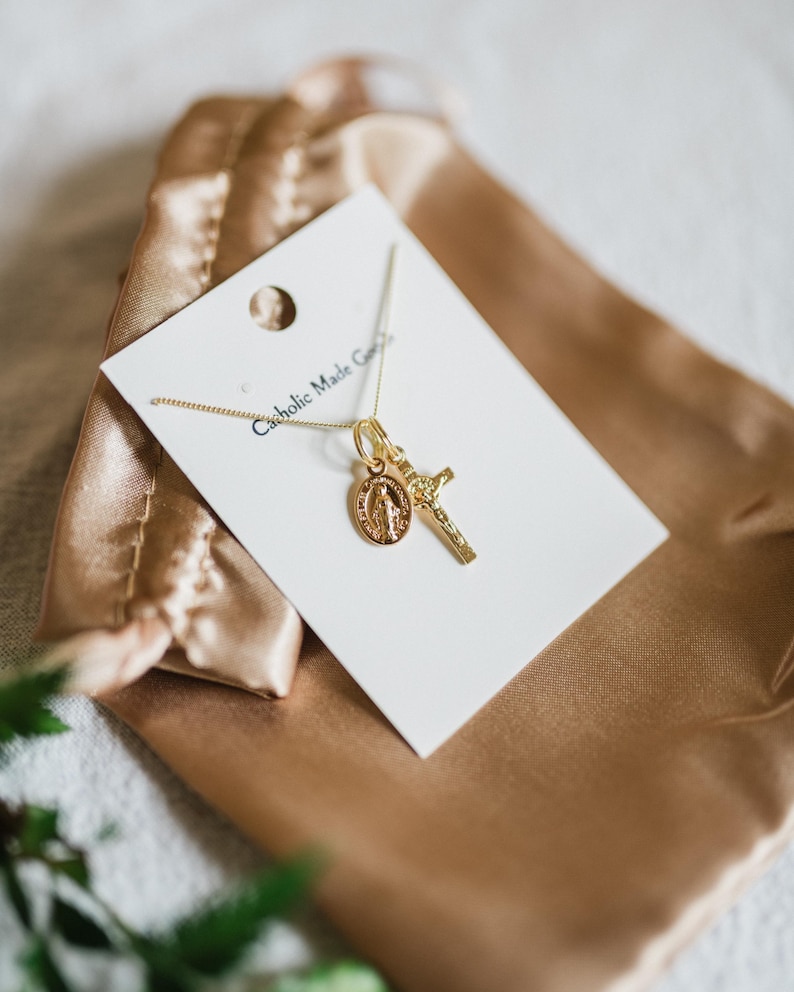 May include: A gold-colored necklace with two pendants: a cross and a medallion featuring a religious figure. The necklace is displayed on a white card inside a gold satin pouch. The card reads "Catholic Made Goods".