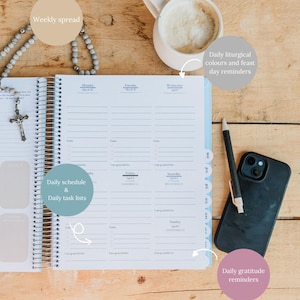 May include: An open weekly planner with a spiral binding, featuring sections for daily schedules, task lists, and gratitude reminders. A cup of coffee, a pen, and a smartphone are next to the planner. A rosary and a cross are also visible.