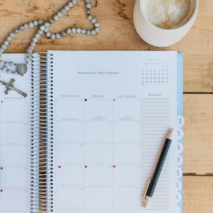 May include: A spiral-bound planner open to May 2020, with the text "Month of the Holy Eucharist." A rosary and a pen rest on the pages. A mug of coffee sits in the upper right corner. The planner has blue tabs.