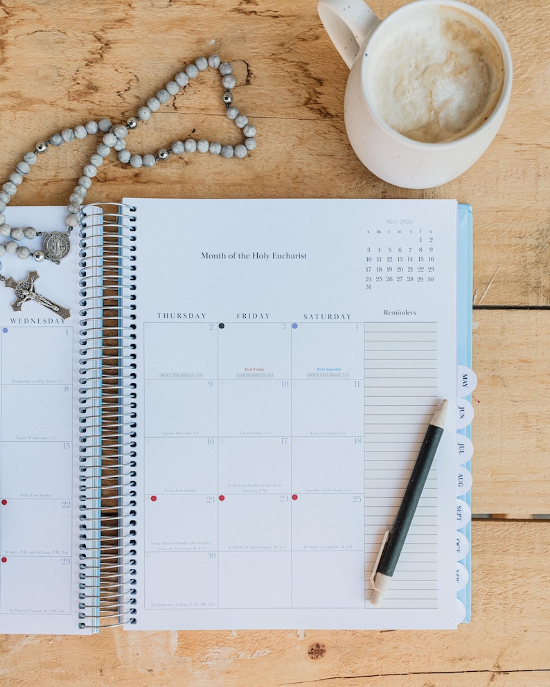 May include: A spiral-bound planner open to May 2020, with the text "Month of the Holy Eucharist." A rosary and a pen rest on the pages. A mug of coffee sits in the upper right corner. The planner has blue tabs.