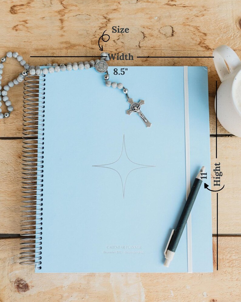 May include: A light blue spiral-bound planner, 8.5 inches wide and 11 inches high, with a silver pen and a rosary. The planner has a star design and the words "CALENDAR PLANNER" printed on it. The background is a light wood surface.