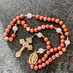 May include: A rosary with coral-colored beads and gold accents. The rosary features a gold crucifix and a gold pendant with an oval image. The beads are interspersed with white and pink beads.