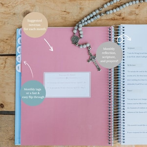 May include: Open pink spiral-bound planner with monthly tabs and a rosary. The planner includes sections for scripture, reflection, and prayers. The cover has text that says "Suggested novenas for each month".