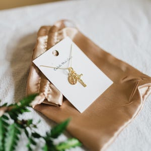 May include: A gold-colored necklace with a pendant featuring a cross and a religious medallion. The necklace is displayed on a white card inside a gold satin pouch. The text "Catholic Made Goods" is printed on the card.
