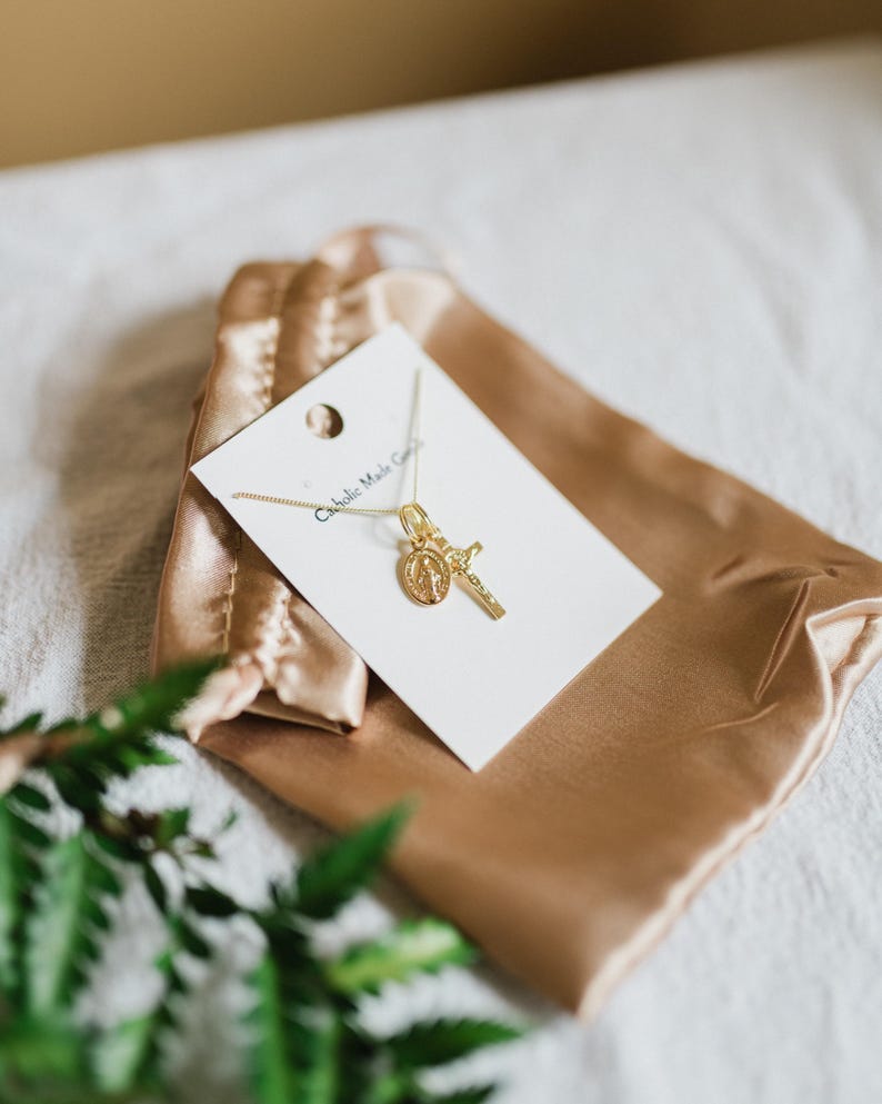 May include: A gold-colored necklace with a pendant featuring a cross and a religious medallion. The necklace is displayed on a white card inside a gold satin pouch. The text "Catholic Made Goods" is printed on the card.
