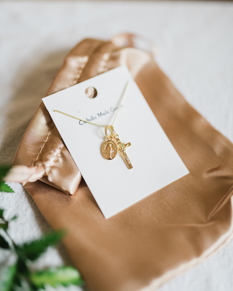 May include: Gold-colored necklace with a pendant featuring a cross and a medallion with a religious figure. The necklace is displayed on a white card with the text "Catholic Made Goods" and rests on a gold satin pouch.