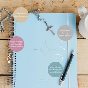 May include: A light blue spiral-bound planner with a matte hard cover, elastic band, and silver metal binding. A rosary and pen rest on the planner. The planner has an inside pocket to hold notes or receipts.