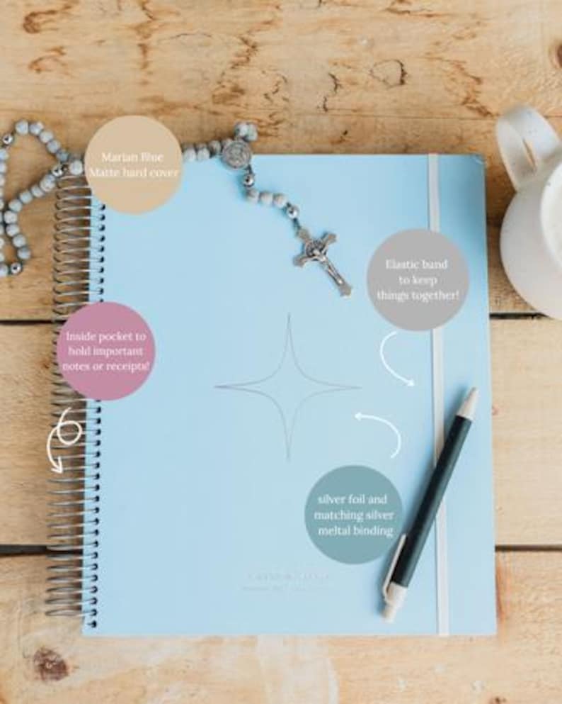 May include: A light blue spiral-bound planner with a matte hard cover, elastic band, and silver metal binding. A rosary and pen rest on the planner. The planner has an inside pocket to hold notes or receipts.
