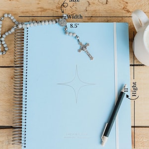 May include: A light blue spiral-bound planner, 8.5 inches wide and 11 inches high, with a silver pen and a rosary. The planner has a star design and the words "CALENDAR PLANNER" printed on it. The background is a light wood surface.