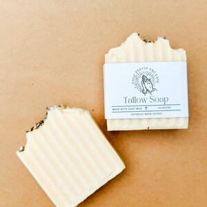 May include: Two bars of Tallow Soap, one partially eaten, on a light brown surface. The soap is off-white with visible lavender flecks. One bar has a white label that reads "Tallow Soap" and "Made with Goat Milk" and "Lavender".