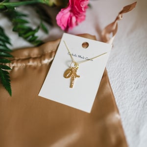 May include: A gold-colored necklace with a cross and a medallion pendant, displayed on a white card. The card has the text "Catholic Made Goods". The necklace rests on a gold satin pouch, with pink flowers and green leaves in the background.
