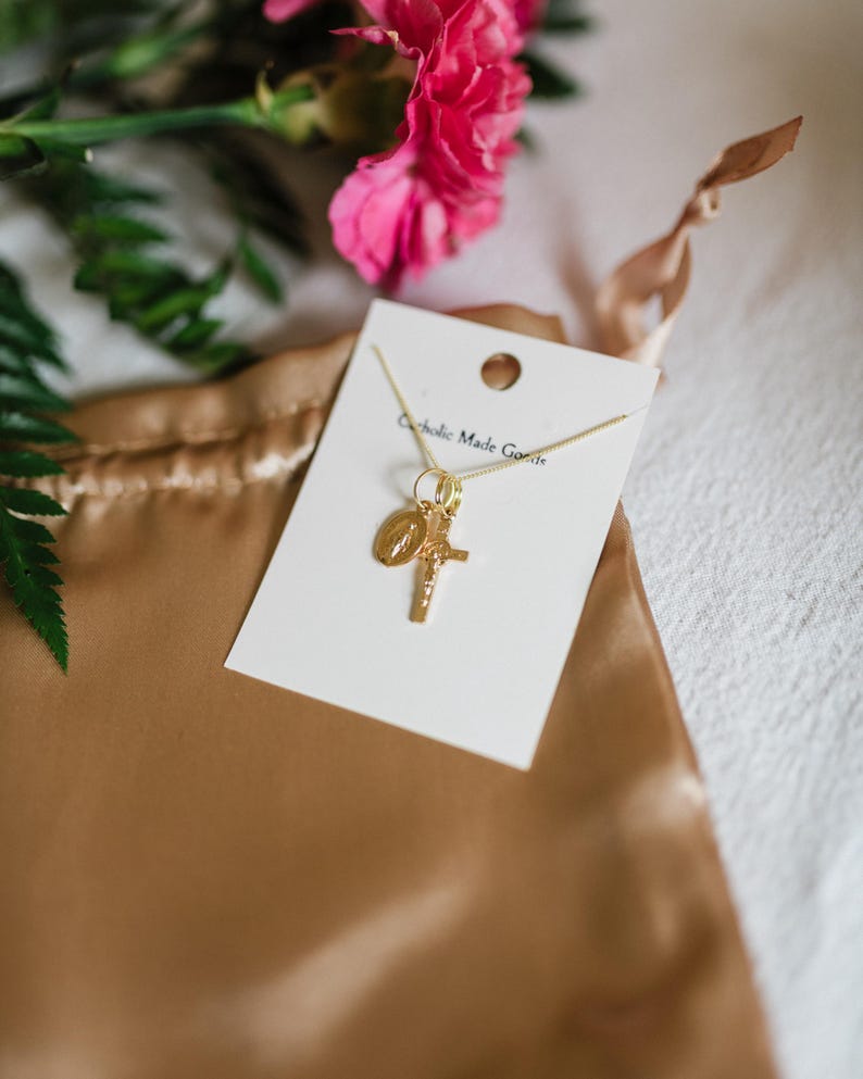 May include: A gold-colored necklace with a cross and a medallion pendant, displayed on a white card. The card has the text "Catholic Made Goods". The necklace rests on a gold satin pouch, with pink flowers and green leaves in the background.
